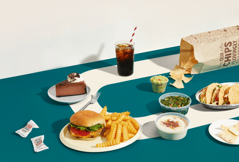 Colorful flatlay of various food items including burger with fries, tacos, chocolate cake, and cold drink with striped straw on teal and white surface