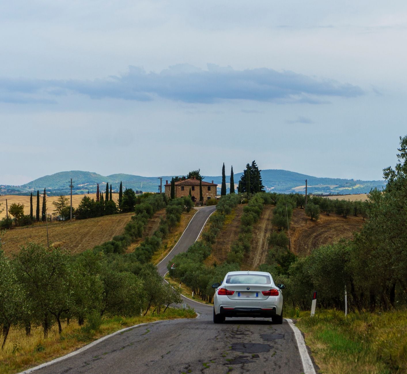 White sedan driving on a winding road through Tuscan olive groves toward a terracotta villa on a hilltop