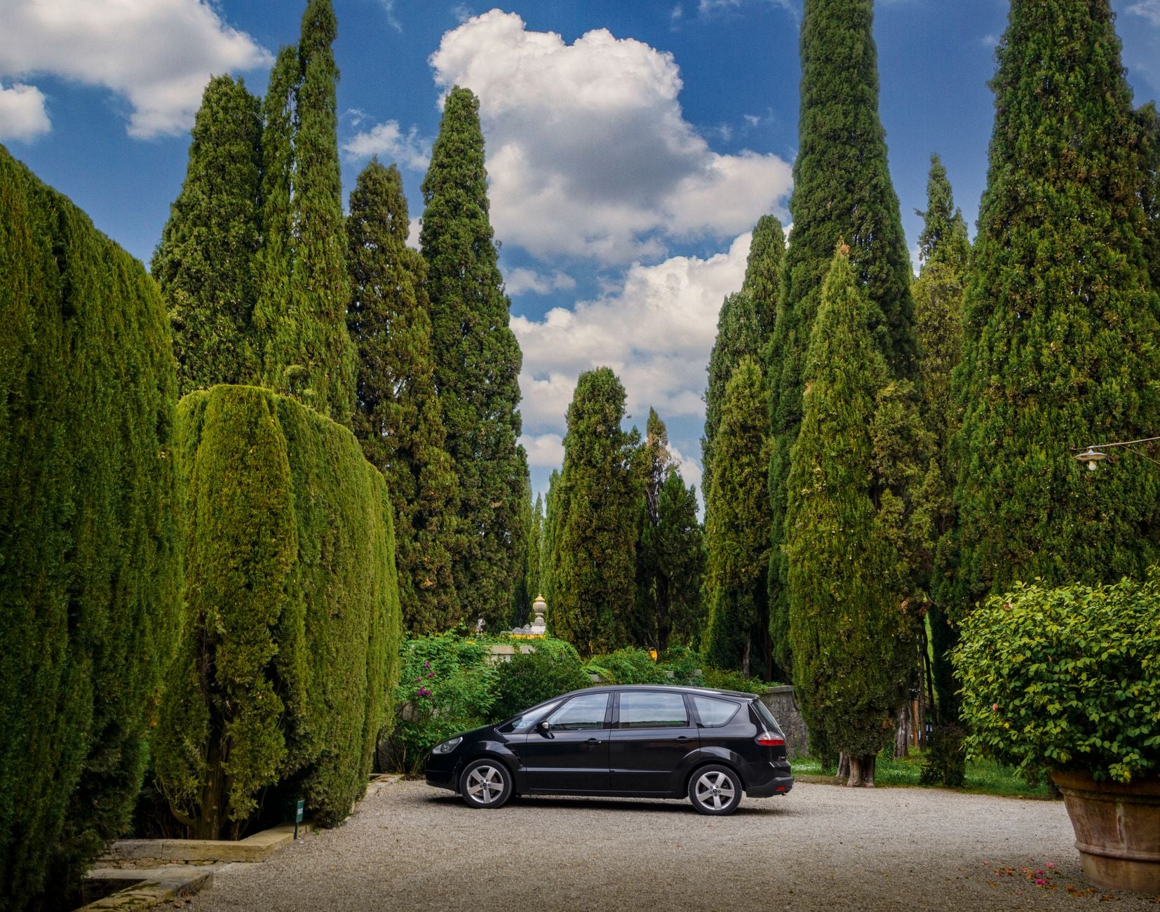 Black SUV parked in a tree-lined cypress driveway with ivy-covered walls and blue sky above