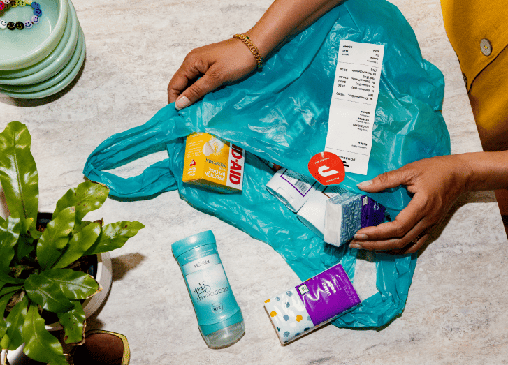 Hands holding open teal reusable shopping bag filled with groceries and household items including milk, snacks, and other products
