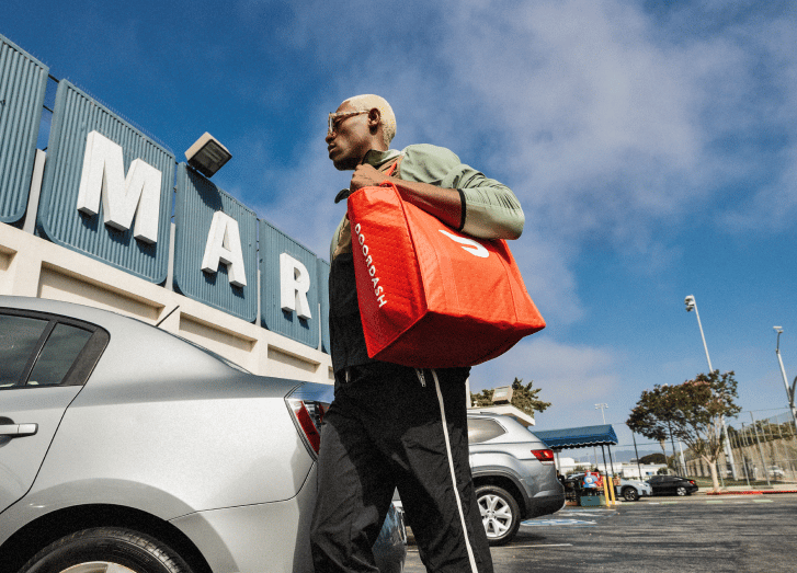 Person carrying a red delivery bag walking in a parking lot near a store